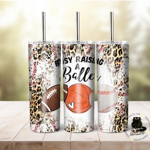 Könnte beinhalten: Drei Edelstahlbecher mit Deckel und Strohhalmen. Die Becher sind mit einem Leopardenmuster, Baseballnähten und einem weißen Hintergrund verziert. Der Text auf den Bechern lautet "Busy Raising a Baller".