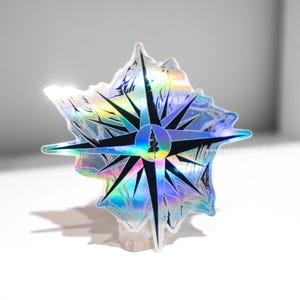 May include: A holographic sticker in the shape of a compass rose. The sticker has a black compass design with a central tree graphic, surrounded by iridescent colors. The sticker is on a white surface.