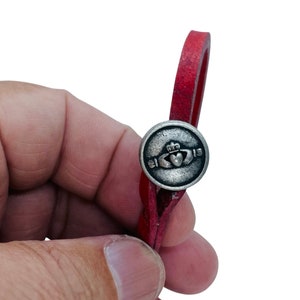 May include: A red leather bracelet with a silver button clasp. The button features a Claddagh design, with a heart, crown, and hands. The bracelet is held against a white background.
