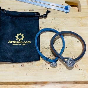 Op de afbeelding: Twee leren armbanden, één blauw en één bruin, met zilverkleurige knoopsluitingen. Een zwart trekkoordzakje met goudkleurige opdruk "Artissun.com Symbols of Light" ligt naast de armbanden. Een liniaal staat op de achtergrond.
