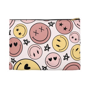 Könnte beinhalten: Eine rosa und gelbe Reißverschlusstasche mit weißem Hintergrund und einem sich wiederholenden Muster aus Smiley-Gesichtern mit verschiedenen Ausdrücken, Sternen und Herzen.