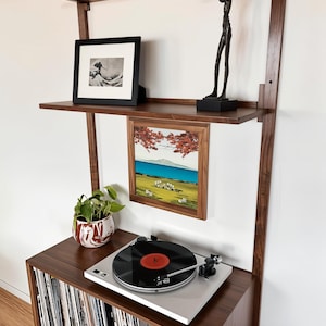 May include: A walnut wood wall-mounted shelf unit with a record player, vinyl records, a potted plant, and framed artwork. The shelf unit has three shelves and a lower cabinet with a record player on top. The artwork includes a black and white photograph and a landscape painting.
