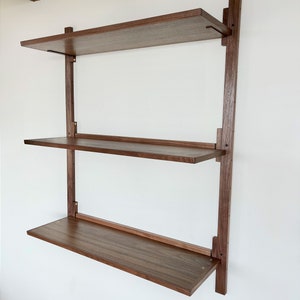May include: A three-tiered wall-mounted shelf unit made of dark brown wood. The shelves are supported by vertical wooden posts and metal brackets. The shelves are all the same width and depth.