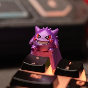 Könnte beinhalten: Ein lila Gengar-Keycap, ein Charakter aus der Pokémon-Serie, sitzt auf einer schwarzen Tastatur. Der Keycap hat ein lächelndes Gesicht mit roten Wangen und einer gezackten Krone. Die Tastaturtasten sind schwarz.