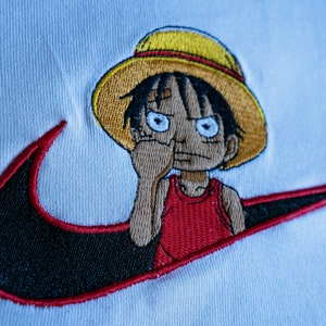 Puede incluir: Camiseta blanca con un swoosh Nike bordado en negro y rojo y un personaje de dibujos animados con un sombrero de paja amarillo y un tanque rojo.