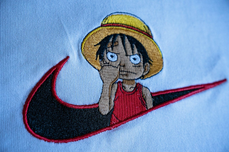 Puede incluir: Camiseta blanca con un swoosh Nike bordado en negro y rojo y un personaje de dibujos animados con un sombrero de paja amarillo y un tanque rojo.