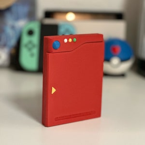Könnte beinhalten: Eine rote Plastik-Replik eines Pokémon-Pokédex. Der Pokédex hat einen blauen Knopf, ein gelbes Dreieck und vier farbige Lichter oben.
