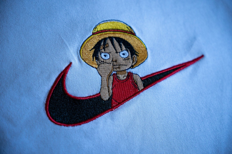 Puede incluir: Camiseta blanca con un swoosh Nike bordado en negro y rojo con un personaje de dibujos animados con un sombrero amarillo y una camisa roja dentro del swoosh.