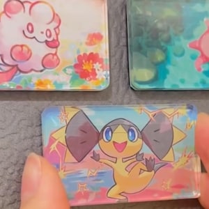 Könnte beinhalten: Rechteckige, transparente Acrylmagnete mit bunten Zeichentrickfiguren. Ein Magnet zeigt eine gelb-braune Figur mit blauen Augen und einem lächelnden Gesichtsausdruck vor einem blau-rosa Hintergrund. Andere Magnete zeigen ähnliche Designs.