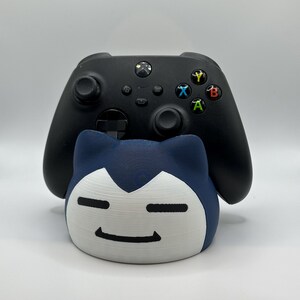 Peut inclure: Une manette Xbox noire est posée au-dessus d'un support en forme de personnage imprimé en 3D bleu et blanc. La manette a des boutons colorés et deux joysticks. Le support a un design de visage souriant.