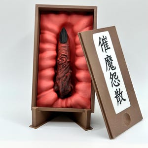 Puede incluir: Una escultura detallada, marrón rojizo, de un dedo con una uña negra, presentada en una caja de exhibición marrón con un interior rojo. La caja tiene una tapa con bisagras con texto japonés.