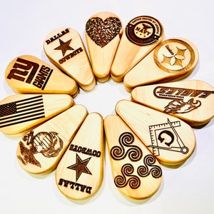 May include: A collection of wooden, teardrop-shaped items with various designs and text. The designs include a star, an American flag, a tree, and logos for the Navy, NFL teams, and a Masonic symbol. The items are arranged in a circular pattern.