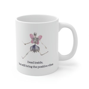 May include: White ceramic mug with a graphic of a skeleton wearing pink fairy wings and the text "Dead inside, but still bring the positive vibes".