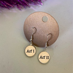 Op de afbeelding: Zilverkleurige oorhangers met ronde hangers. Elke hanger is crèmekleurig en beschreven met "Act I" en "Act II" in zwart schrift. De oorbellen worden gepresenteerd tegen een lichtbruine achtergrond, met een paars decoratief element.