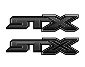 May include: Two black STX logo decals. Each decal features the letters "STX" in a bold, blocky font, followed by a stylized "X" shape. The design has a metallic, three-dimensional appearance with a dark gray gradient.