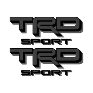 Op de afbeelding: Twee zwart-grijze TRD Sport logo's, elk met een 3D-effect, worden op een witte achtergrond weergegeven. De tekst is vet en gestileerd, met het woord "SPORT" onder de letters "TRD".