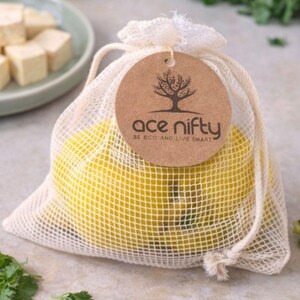 May include: A mesh bag filled with yellow lemons, tied with a drawstring. A brown tag with the text "ace nifty" and a tree logo hangs from the bag. The bag is on a light-colored surface with green herbs and a plate of tofu cubes in the background.