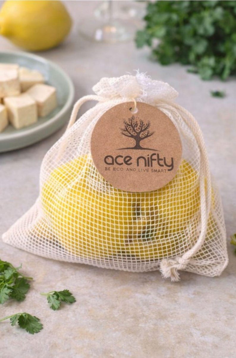 May include: A mesh bag filled with yellow lemons, tied with a drawstring. A brown tag with the text "ace nifty" and a tree logo hangs from the bag. The bag is on a light-colored surface with green herbs and a plate of tofu cubes in the background.