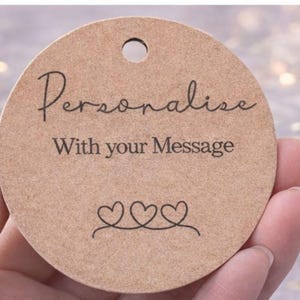 May include: A round, light brown tag with a hole at the top. The tag features the words "Personalise With your Message" in black script and a line of three connected heart shapes. The tag is held in a person's hand.