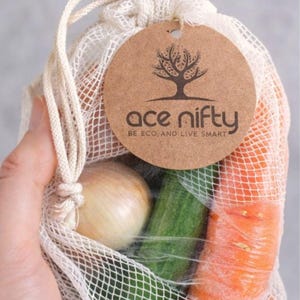 May include: A reusable mesh produce bag filled with fresh vegetables, including a carrot, cucumber, and onion. The bag has a drawstring closure and a round tag with the text "ace nifty BE ECO. AND LIVE SMART".