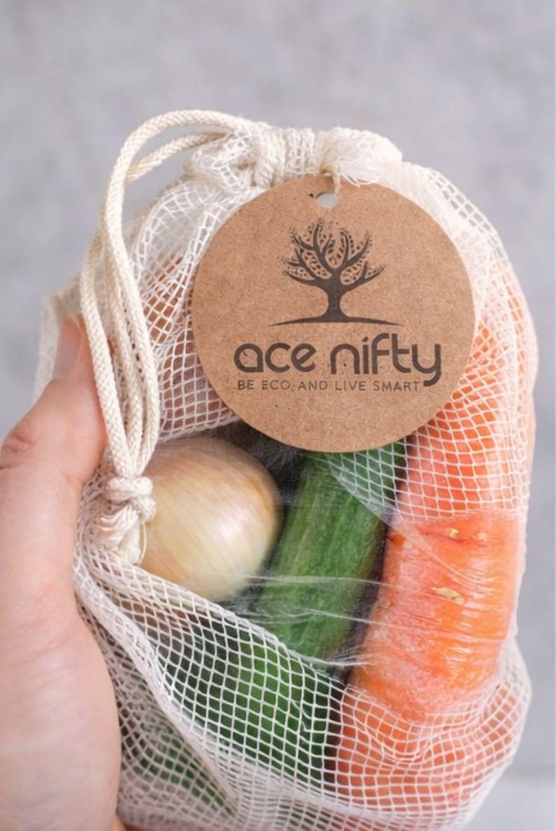 May include: A reusable mesh produce bag filled with fresh vegetables, including a carrot, cucumber, and onion. The bag has a drawstring closure and a round tag with the text "ace nifty BE ECO. AND LIVE SMART".