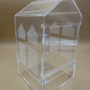 Könnte beinhalten: Ein transparentes Acryl-Terrarium in Hausform. Die Struktur verfügt über eine Scharniertür, Belüftungslöcher und ein Logo auf der Basis. Das Design umfasst ein Satteldach und rechteckige Fenster, geeignet für kleine Haustiere.