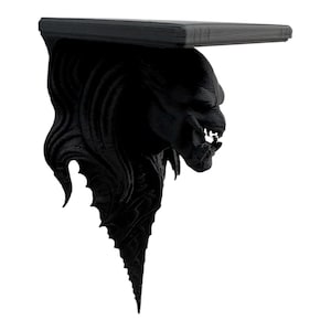 May include: A black, 3D-printed shelf featuring a detailed, monstrous face as a support. The shelf has a flat top and a textured, stylized design. The shelf is a unique decorative item.