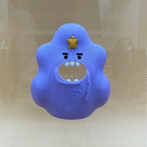 May include: A whimsical, lavender-colored, blob-shaped figurine with a wide open mouth and small white teeth. The figurine has a gold star-shaped badge on its head and two small black eyes. The design has a textured, layered appearance.