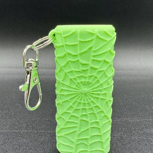 May include: A bright green silicone keychain with a spiderweb design. The keychain has a silver metal ring and a silver metal clasp.