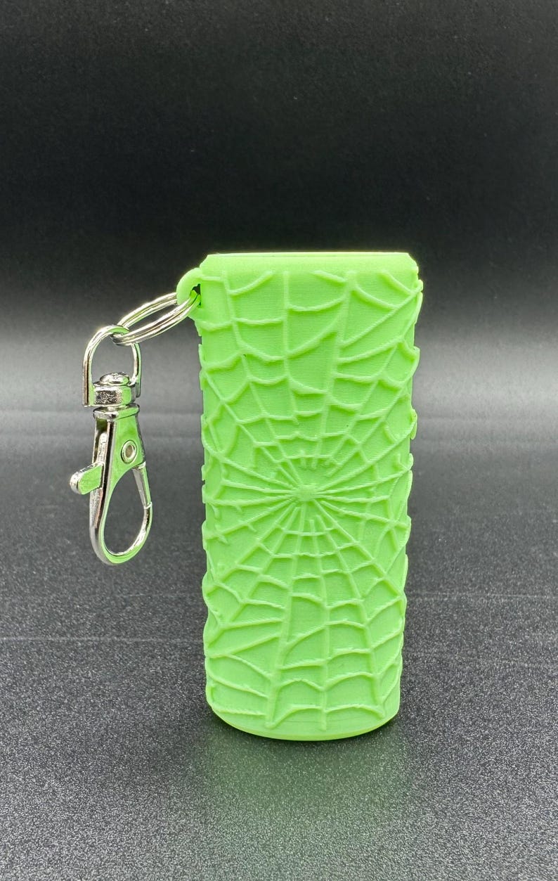 May include: A bright green silicone keychain with a spiderweb design. The keychain has a silver metal ring and a silver metal clasp.