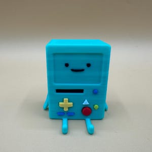 May include: A turquoise, rectangular figurine with a smiling face, black eyes, and a black horizontal line for a mouth. It has a yellow plus-shaped button, a red button, and blue and green buttons. The figurine has small arms and legs.