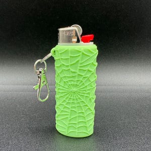 May include: A green lighter with a spiderweb design and a silver keychain attached. The lighter has a red button and the text "BIC" is visible on the side.