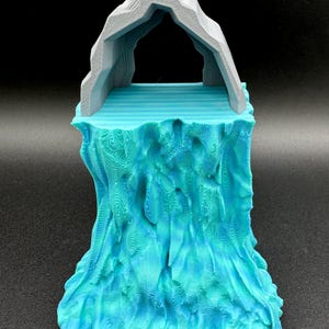 Pode incluir: Uma cena em miniatura impressa em 3D com um arco de rocha cinza sobre uma base de água azul turquesa. A base tem uma superfície texturizada que se assemelha a ondas.