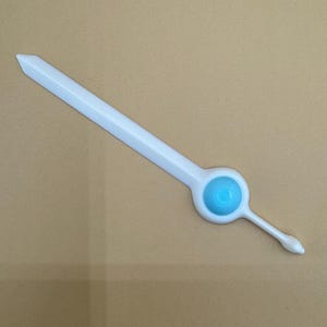 May include: A white plastic sword-shaped toy with a light blue circular design on the handle. The toy has a pointed tip and a long, straight blade. The handle has a small, thin extension.