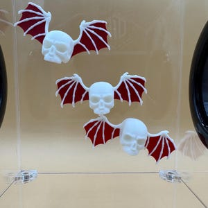 May include: Three white skull-shaped ornaments with red bat wings are displayed. The skulls have detailed facial features. The wings are a deep red color. The ornaments are arranged in a vertical line against a clear background.