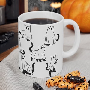 May include: White ceramic mug with a black and white illustration of cats dressed as ghosts. The mug has a black handle and is filled with a dark liquid.