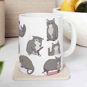 Op de afbeelding: Witte keramische mok met een grijs kattenontwerp. De kat is in verschillende poses, waaronder zittend, slapend en er boos uitziend. De mok heeft een handvat aan de rechterkant.