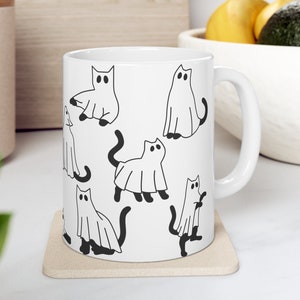 May include: White ceramic mug with a black and white illustration of cats dressed as ghosts. The mug has a handle and is sitting on a beige coaster.