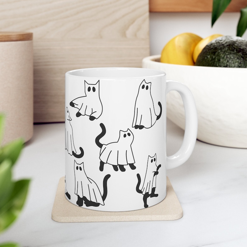 May include: White ceramic mug with a black and white illustration of cats dressed as ghosts. The mug has a handle and is sitting on a beige coaster.