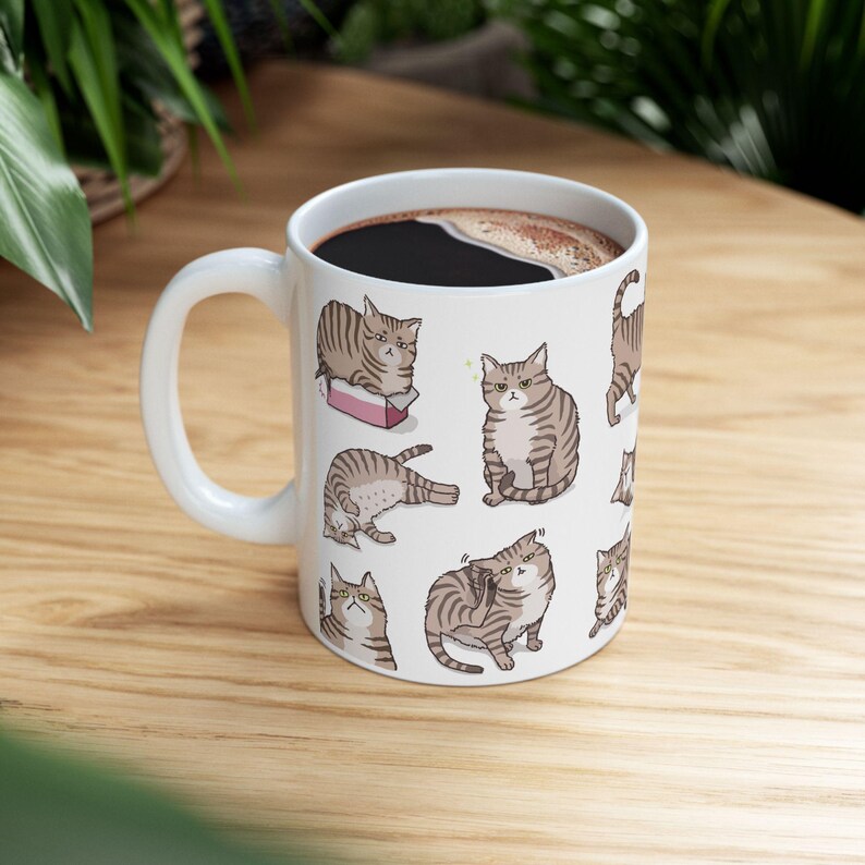 May include: White ceramic mug with a brown tabby cat design. The cat is shown in various poses, including sleeping, sitting, and looking at the camera. The mug is filled with coffee.