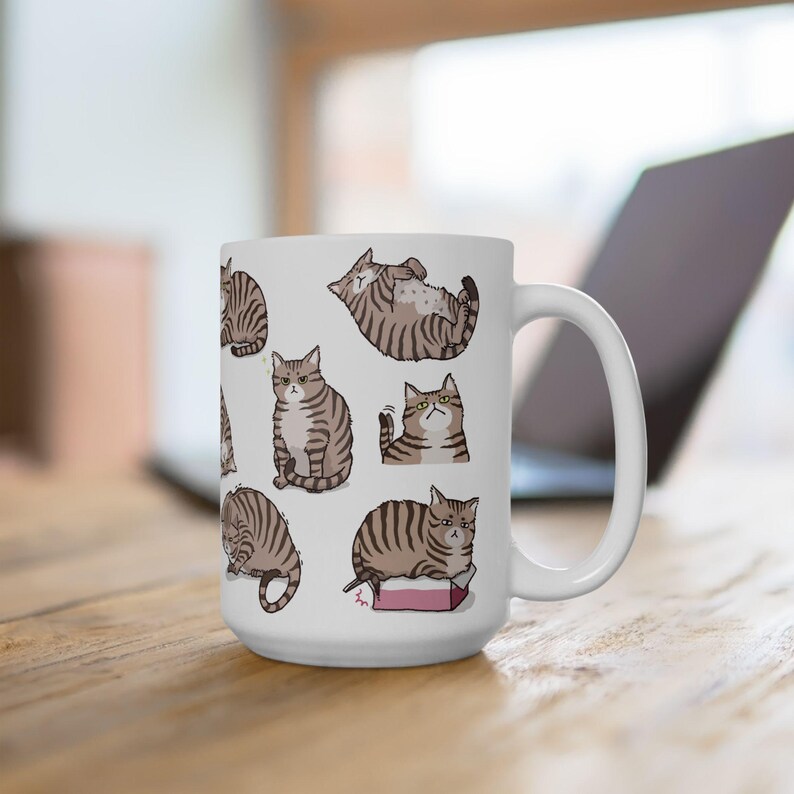 May include: White ceramic mug with a pattern of brown tabby cats in various poses. The cats are illustrated in a cartoon style.
