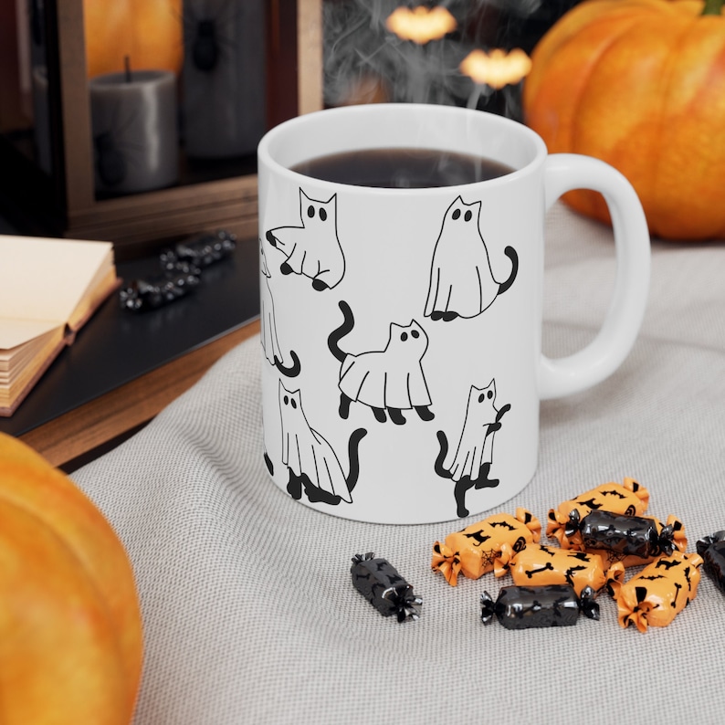 May include: White ceramic mug with a black and white illustration of cats dressed as ghosts. The mug has a black handle and is filled with a dark liquid.