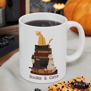 May include: White ceramic mug with a handle, filled with a dark liquid. The mug features a design of three cats sitting on a stack of books, with the text "Books & Cats" below. A pumpkin and candy are in the background.