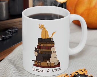 May include: White ceramic mug with a handle, filled with a dark liquid. The mug features a design of three cats sitting on a stack of books, with the text "Books & Cats" below. A pumpkin and candy are in the background.