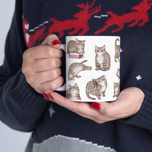 May include: White ceramic mug with a design of multiple brown tabby cats in various poses. The cats are all cartoon-style and have different expressions.