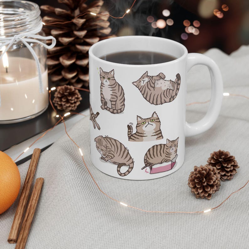 May include: White ceramic mug with a black handle featuring a repeating pattern of brown tabby cats in various poses. The mug is filled with a dark liquid.
