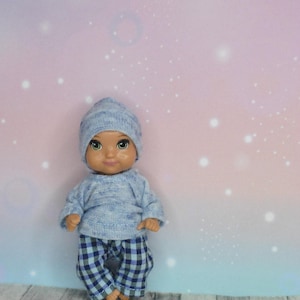 Könnte beinhalten: Eine kleine Puppe, die einen blau-weiß karierten Schlafanzug mit einer passenden blauen Strickmütze trägt. Die Puppe hat braune Augen und einen hellbraunen Teint.