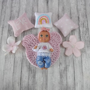 以下が含まれることがあります： 白い花柄のシャツを着て、青いジーンズを履き、ピンクの編み物帽子をかぶった人形。人形はピンクの編み物毛布の上に横たわっており、両側にピンクの花が置かれている。背景には刺繍模様の入ったピンクの枕が3つある。