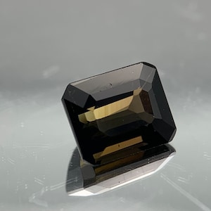 May include: A dark brown emerald cut gemstone with a slight golden hue.