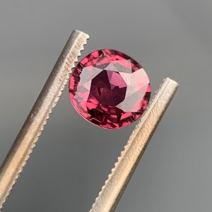 May include: A round, faceted, deep red gemstone held in tweezers. The gemstone is a rich, dark red colour with a slight purple hue.